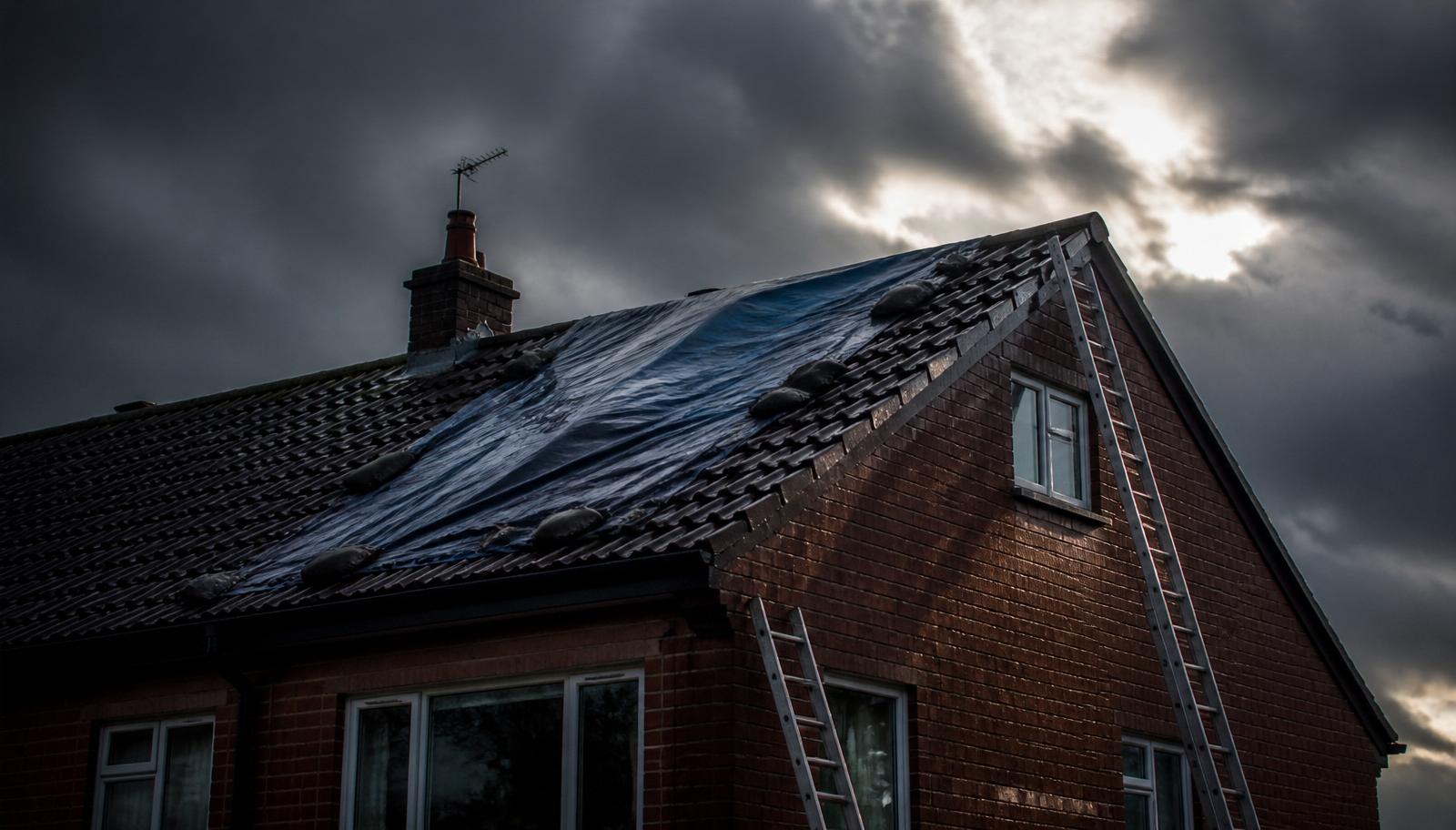 Storm-damaged Yorkshire tiled roof covered with weighted tarpaulin sheeting and ladders in place, ready for emergency roofing repair